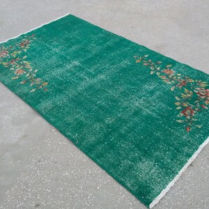 Pode incluir: Um tapete verde vintage com um padr&atilde;o floral em tons de laranja e amarelo. O tapete tem uma borda de franjas brancas.