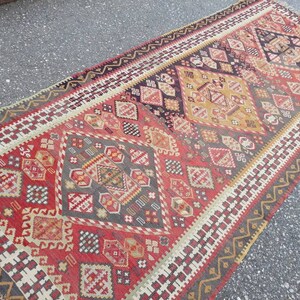 May include: A vintage Turkish rug with a geometric pattern in shades of red, white, black, and gold. The rug has a fringed edge and is woven with intricate details.