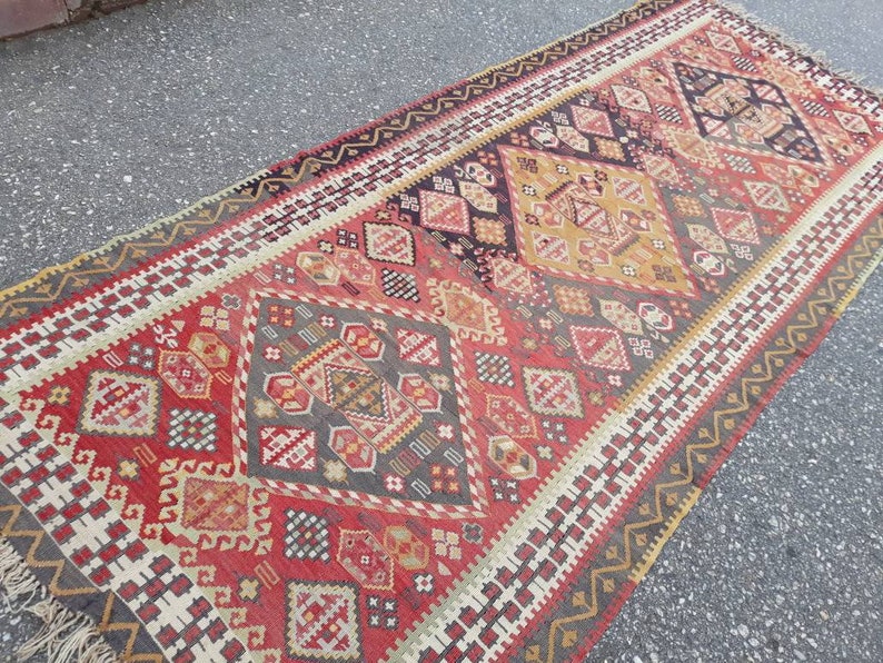 May include: A vintage Turkish rug with a geometric pattern in shades of red, white, black, and gold. The rug has a fringed edge and is woven with intricate details.