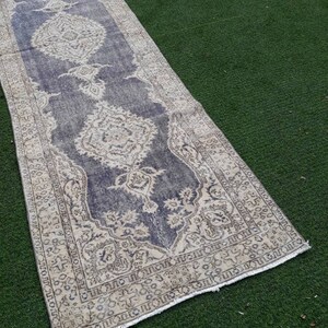 May include: A vintage runner rug with a faded blue background and a cream-colored floral pattern. The rug has a frayed edge and is laid out on a green surface.