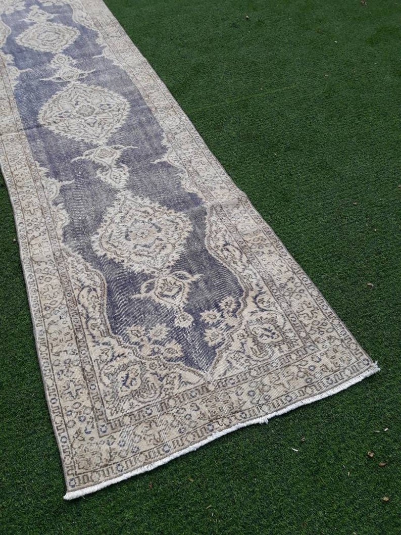 May include: A vintage runner rug with a faded blue background and a cream-colored floral pattern. The rug has a frayed edge and is laid out on a green surface.