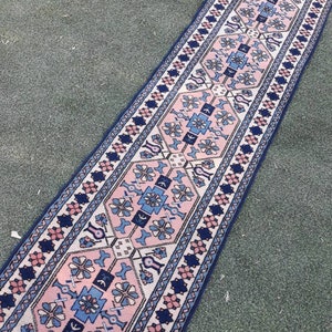 May include: A long, narrow rug with a pink, blue, and white geometric pattern. The rug has a traditional design with intricate details.