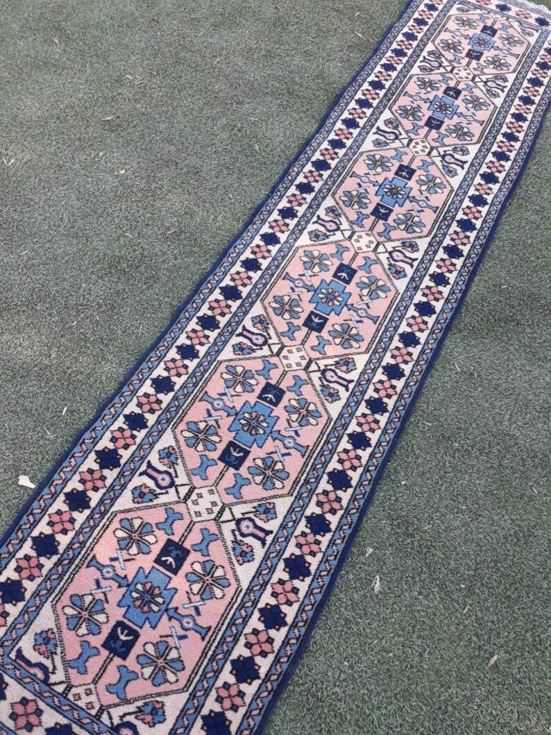May include: A long, narrow rug with a pink, blue, and white geometric pattern. The rug has a traditional design with intricate details.