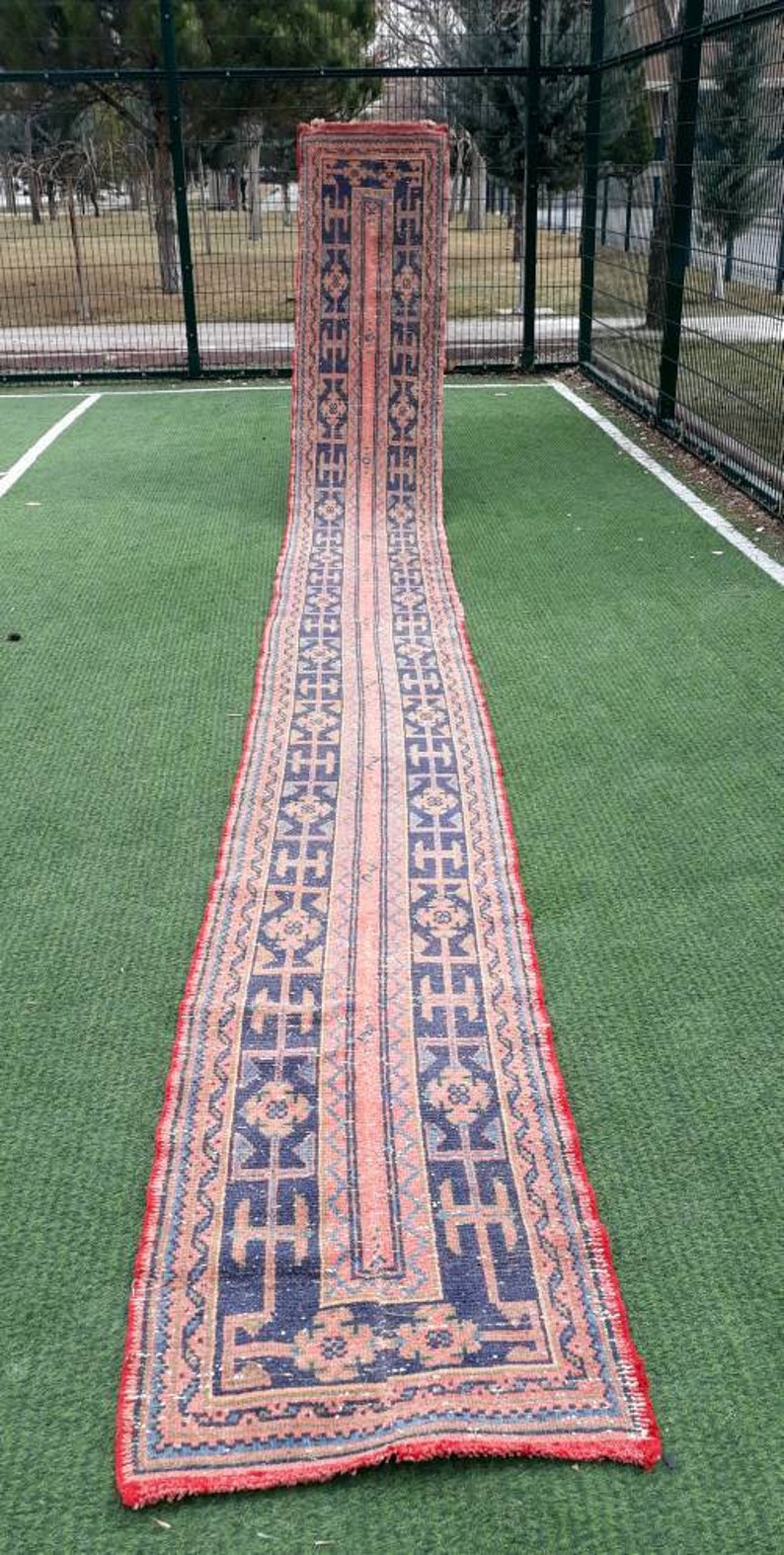 May include: A long, narrow rug with a traditional Persian design. The rug has a red border with a blue and pink center. The center of the rug features a geometric pattern.