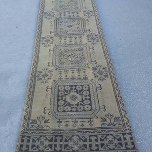 May include: A long, rectangular rug with a beige background and a black geometric pattern. The rug has a fringe border.