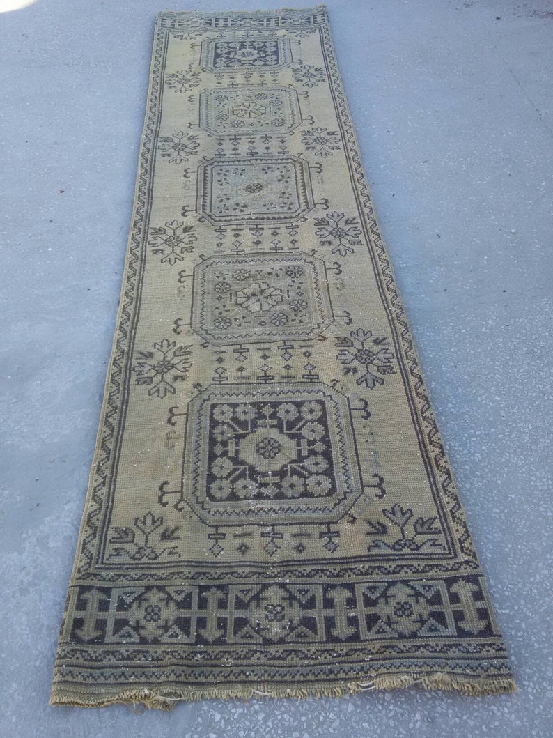 May include: A long, rectangular rug with a beige background and a black geometric pattern. The rug has a fringe border.