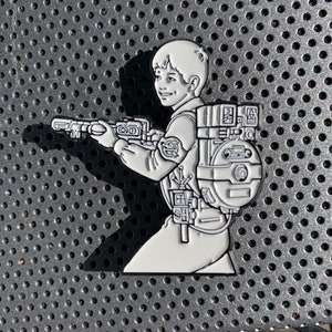 May include: White enamel pin of a cartoon boy wearing a backpack and holding a gun. The boy is smiling and has short brown hair. The backpack has a lot of detail and is made up of many different compartments.