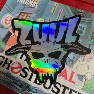 May include: A silver holographic sticker with the word "ZUL" in black letters and a detailed illustration of a monster with horns and fangs. The sticker is on a background with the words "Ghostbusters" and "Aura".