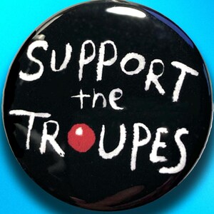 May include: A round, black button with the words "SUPPORT the TROUPES" in white, handwritten-style lettering. A red circle is used as the "O" in TROUPES. The text "pencilisland" is written in pink at the bottom.