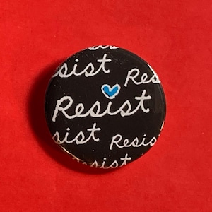 May include: Black button with white text that says "Resist" with a blue heart in the middle.