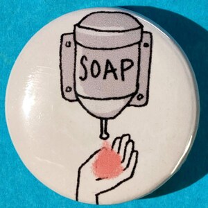 May include: A white button with a cartoon illustration of a hand receiving soap from a dispenser labeled "SOAP".