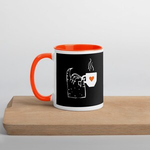 Könnte beinhalten: Weißer Keramikbecher mit orangefarbenem Henkel und Innenraum. Der Becher hat einen schwarzen Hintergrund mit einer weißen Illustration einer Hand, die eine Kaffeetasse mit einem Herz hält. Der Becher steht auf einer Holzoberfläche.