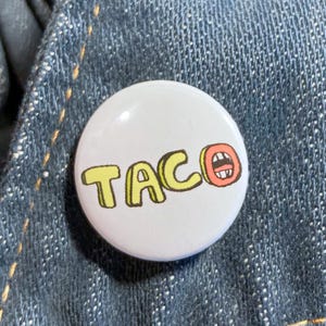May include: A white button with the word "TACO" in yellow letters with a brown outline. The "O" in "TACO" is a cartoon taco with a red mouth and white teeth. The button is on a blue denim background.