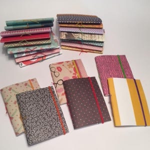 May include: A collection of small, handmade notebooks with various patterned covers. The notebooks are bound with colourful string and have elastic bands to keep them closed. The covers feature patterns such as polka dots, stripes, and floral designs.