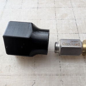 May include: A black, geometric-shaped connector next to a metal fitting with "GasGuard" printed on it. The fitting has a brass-colored hexagonal end. The items are on a light-colored wooden surface.