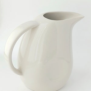 May include: A cream-coloured ceramic jug with a curved handle and a spout for pouring. The jug has a smooth, glossy finish and a rounded body. It is designed for serving beverages.