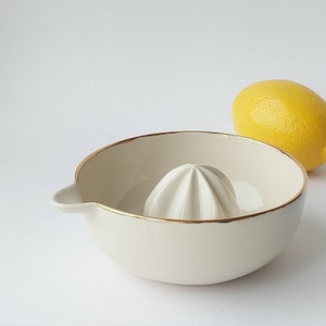 May include: Cream-colored ceramic citrus juicer with a gold rim and a built-in reamer. A yellow lemon rests beside the juicer. The juicer has a spout for easy pouring. Kitchenware.