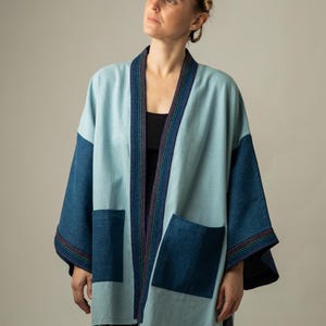 Op de afbeelding: Een lichtblauw jasje met open voorzijde, donkerblauwe mouwen en twee grote donkerblauwe opgestikte zakken. Het kledingstuk is afgewerkt met een donkerblauwe bies langs de kraag en mouwboorden, voorzien van dunne, veelkleurige verticale strepen.