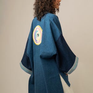 Op de afbeelding: Een blauwe spijkerjas met wijde mouwen en zijsplitten. De achterkant heeft een cirkelvormige geborduurde patch met een centraal yin-yang symbool omringd door concentrische ringen in geel, oranje, roze en blauw. De mouwboorden hebben gestreepte biezen.
