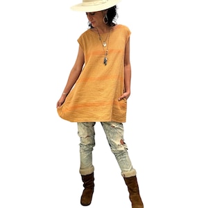 May include: A woman wearing a loose-fitting, sleeveless, orange and yellow striped tunic top with a white hat and distressed blue jeans. She is wearing brown boots.