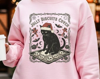 Op de afbeelding: Roze sweatshirt met een afbeelding van een zwarte kat met een kerstmuts, die schedelvormige koekjes bakt. De tekst "JOLLY BISCUITS COVEN" staat boven de kat. Het model draagt een geruite rok en houdt een witte koffiebeker vast.