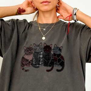 May include: Dark gray t-shirt featuring an embroidered design of four stylized cats. Each cat has a unique pattern, including plaid and patchwork designs. The word "PEPPER" is printed at the bottom right.