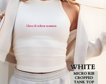 May include: White cropped tank top with a micro rib texture. The tank top has the text "i love it when women" printed on the front in pink.