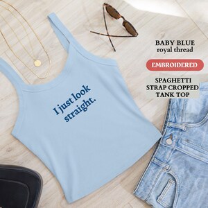 Puede incluir: Una camiseta sin mangas corta azul bebé con tirantes finos con el texto "I just look straight". La camiseta se combina con pantalones cortos de mezclilla, un collar dorado, gafas de sol y un bolso negro. La camiseta está bordada.