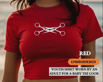 Può includere: Maglietta rossa di taglia bambino con un design ricamato bianco di forbici incrociate. Il testo "RED EMBROIDERED" e "YOUTH SHIRT WORN BY AN ADULT FOR A BABY TEE LOOK" è stampato sulla maglietta.