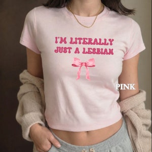 以下が含まれることがあります： レトロなフォントで「I'm literally just a lesbian」と書かれたピンクのTシャツ。テキストの下にピンクのリボンがあります。