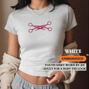 May include: White, embroidered youth shirt featuring a pink crossed scissors design. The shirt is worn by an adult for a baby tee look. The text "WHITE" and "EMBROIDERED" are displayed on the shirt.