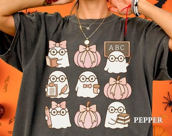 Puede incluir: Camiseta gris oscuro con un patrón repetido de fantasmas de dibujos animados con gafas y lazos, con acentos de calabaza rosa. Algunos fantasmas sostienen libros, lápices y una pizarra con "ABC". La palabra "PEPPER" está impresa.