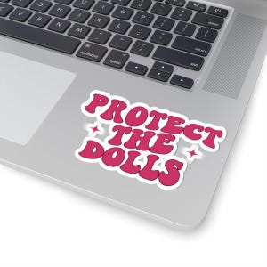May include: A pink sticker with the text "PROTECT THE DOLLS" in a retro font, with a white outline and star accents. The sticker is placed on a laptop. The laptop is silver in colour.