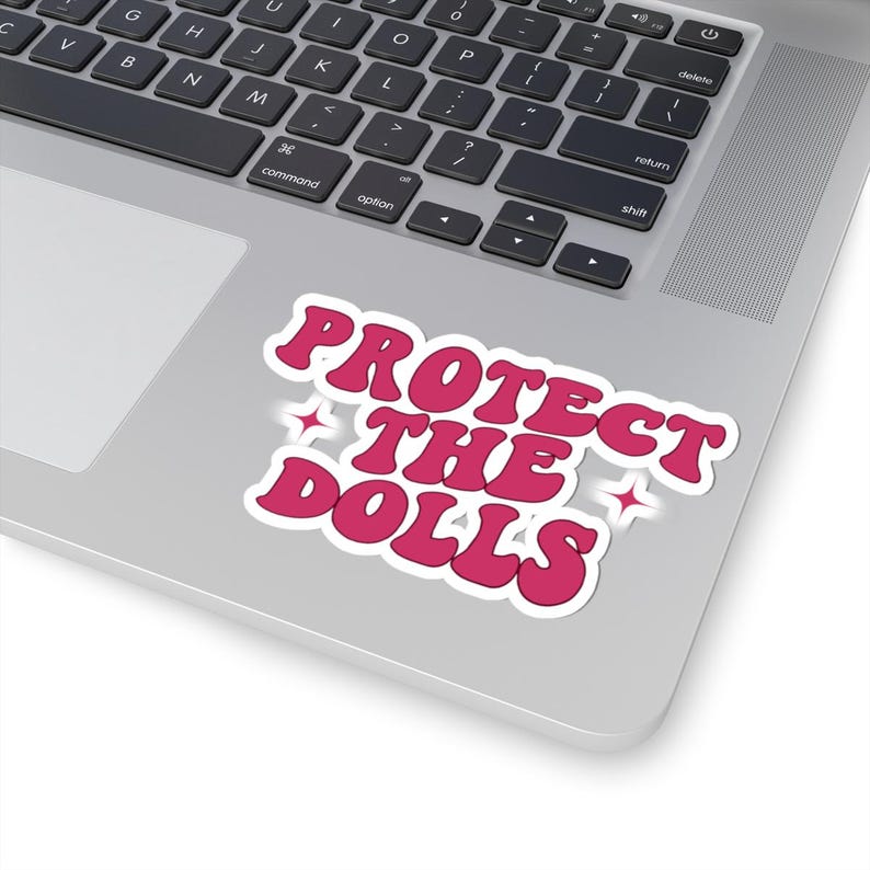May include: A pink sticker with the text "PROTECT THE DOLLS" in a retro font, with a white outline and star accents. The sticker is placed on a laptop. The laptop is silver in colour.