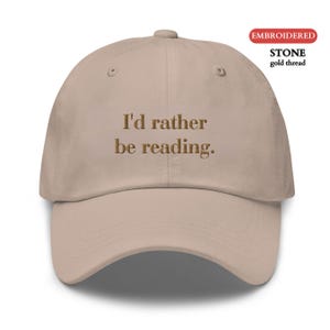 Peut inclure: Casquette de baseball beige avec la phrase brodée "I'd rather be reading." Le texte est en fil d'or. La casquette est en pierre.