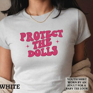 May include: White t-shirt featuring the pink text "PROTECT THE DOLLS" with star details. The shirt is described as a youth shirt worn by an adult for a baby tee look.