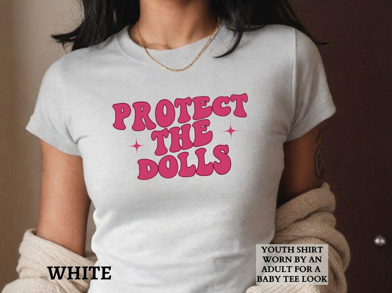 May include: White t-shirt featuring the pink text "PROTECT THE DOLLS" with star details. The shirt is described as a youth shirt worn by an adult for a baby tee look.