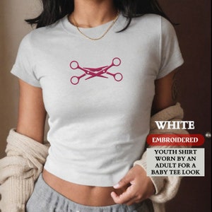 May include: White youth shirt with a pink embroidered design of crossed scissors. The shirt is described as "Youth shirt worn by an adult for a baby tee look."