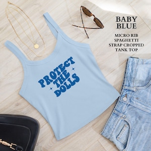 Puede incluir: Un top corto de tirantes finos de micro-canalé azul bebé con el texto "PROTECT THE DOLLS" en azul. El top se combina con pantalones cortos vaqueros, gafas de sol y un collar dorado.