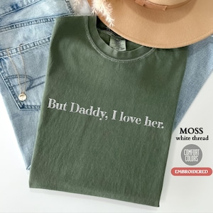 Puede incluir: Una camiseta verde con el texto "But Daddy, I love her." bordado en hilo blanco. La camiseta está hecha de una tela suave y cómoda y es perfecta para el uso diario.