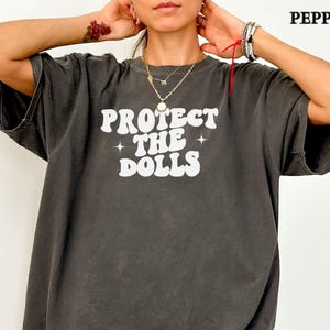May include: Dark grey oversized t-shirt featuring the white text "PROTECT THE DOLLS" in a retro font. The shirt has a faded, vintage appearance. This is a casual, comfortable style of t-shirt.