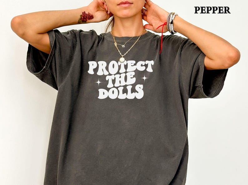 May include: Dark grey oversized t-shirt featuring the white text "PROTECT THE DOLLS" in a retro font. The shirt has a faded, vintage appearance. This is a casual, comfortable style of t-shirt.