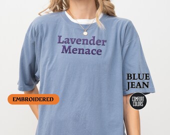 Può includere: Una t-shirt blu jeans con il testo "Lavender Menace" ricamato in viola sul davanti. La t-shirt è descritta come "ricamata" e "colori comfort" nell'immagine.