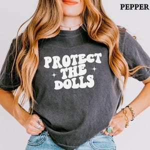May include: Dark grey t-shirt with the text "PROTECT THE DOLLS" in white, retro-style lettering. The shirt has a vintage, worn look. The word "PEPPER" is in the top right corner.