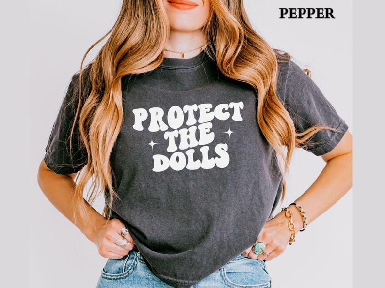 May include: Dark grey t-shirt with the text "PROTECT THE DOLLS" in white, retro-style lettering. The shirt has a vintage, worn look. The word "PEPPER" is in the top right corner.