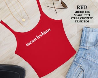 May include: A red micro rib spaghetti strap cropped tank top with the text "mean lesbian" in white. The top is paired with denim shorts, sunglasses, and a gold necklace. The top is a stylish piece of apparel.