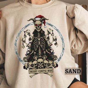 May include: Sand-colored sweatshirt featuring a skeletal figure with horns wearing a Santa hat, surrounded by a barbed wire circle. The skeleton is adorned with a Christmas tree design and a banner reading "MERRY CORPSE-MAS".