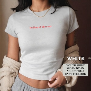 Könnte beinhalten: Ein hellgraues Jugend-T-Shirt mit dem roten Text "lesbian of the year". Das Shirt wird von einem Erwachsenen für einen Baby-T-Shirt-Look getragen. Das Wort "WHITE" ist ebenfalls sichtbar.