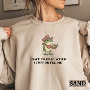 May include: Sand-coloured sweatshirt with a cartoon frog in a cowboy hat reading a book. The text "I HAVE TO READ WEIRD STUFF OR I'LL DIE" is printed below the frog. The word "SAND" is printed at the bottom of the sweatshirt.