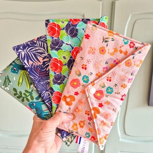 Peut inclure: Quatre pochettes rectangulaires en tissu avec fermetures à pression, présentant divers motifs floraux et botaniques dans des tons de rose, vert, bleu et orange. Une pochette a un fond rose clair avec des petites fleurs et une pression blanche, avec une étiquette 1-2-3 PICOTI.